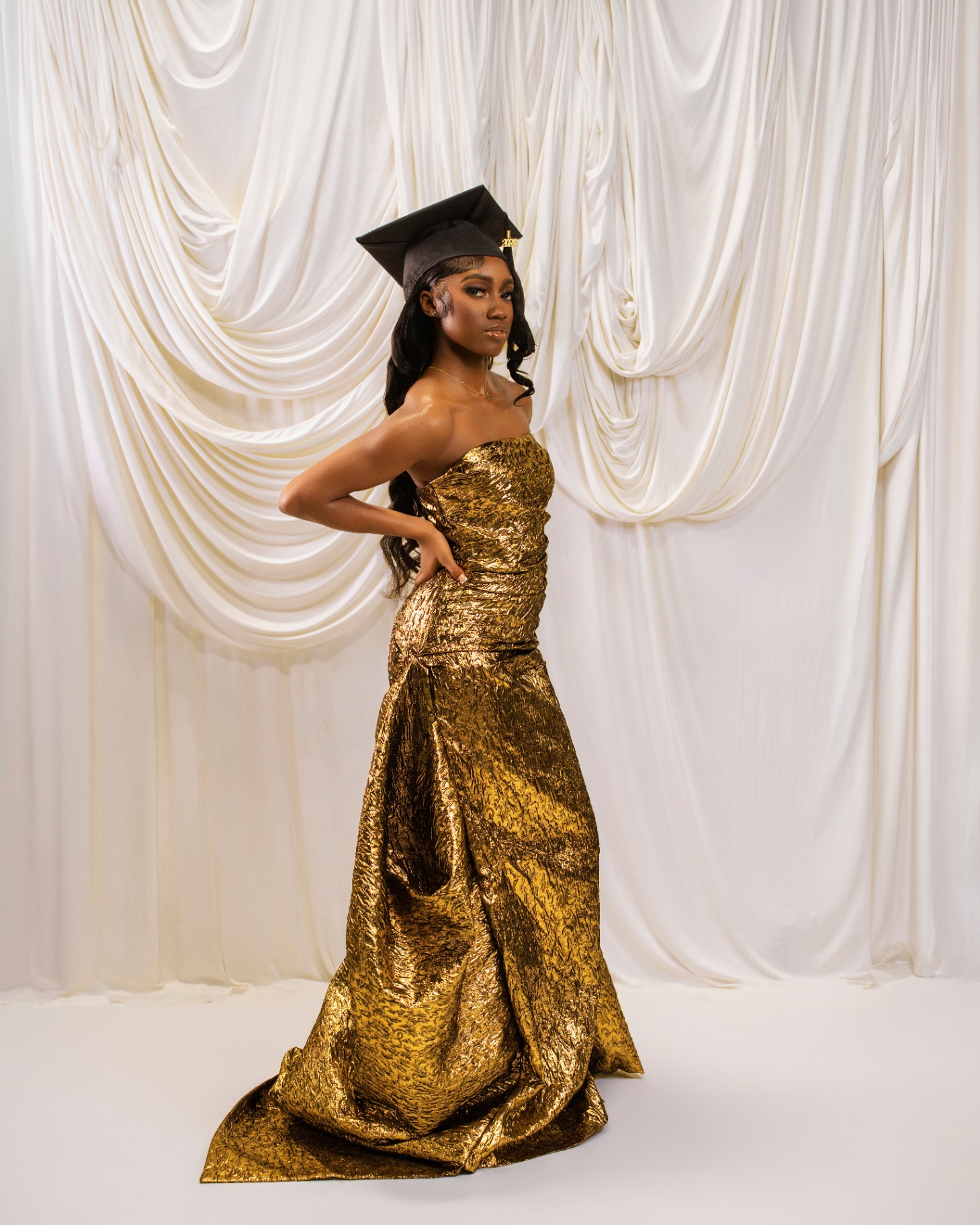 Grad in gold mermaid gown with cap on ivory drapes