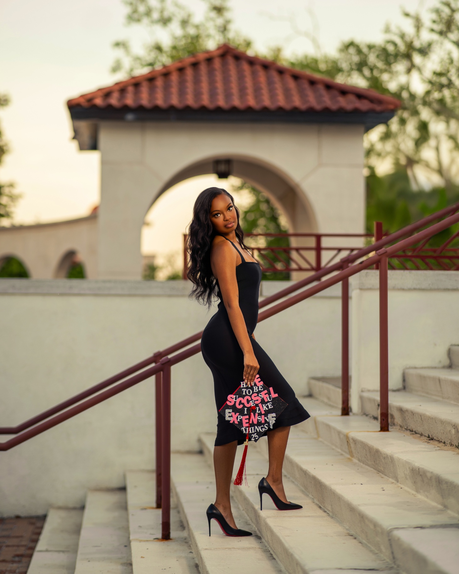 Grad in black dress holding decorated cap at sunset