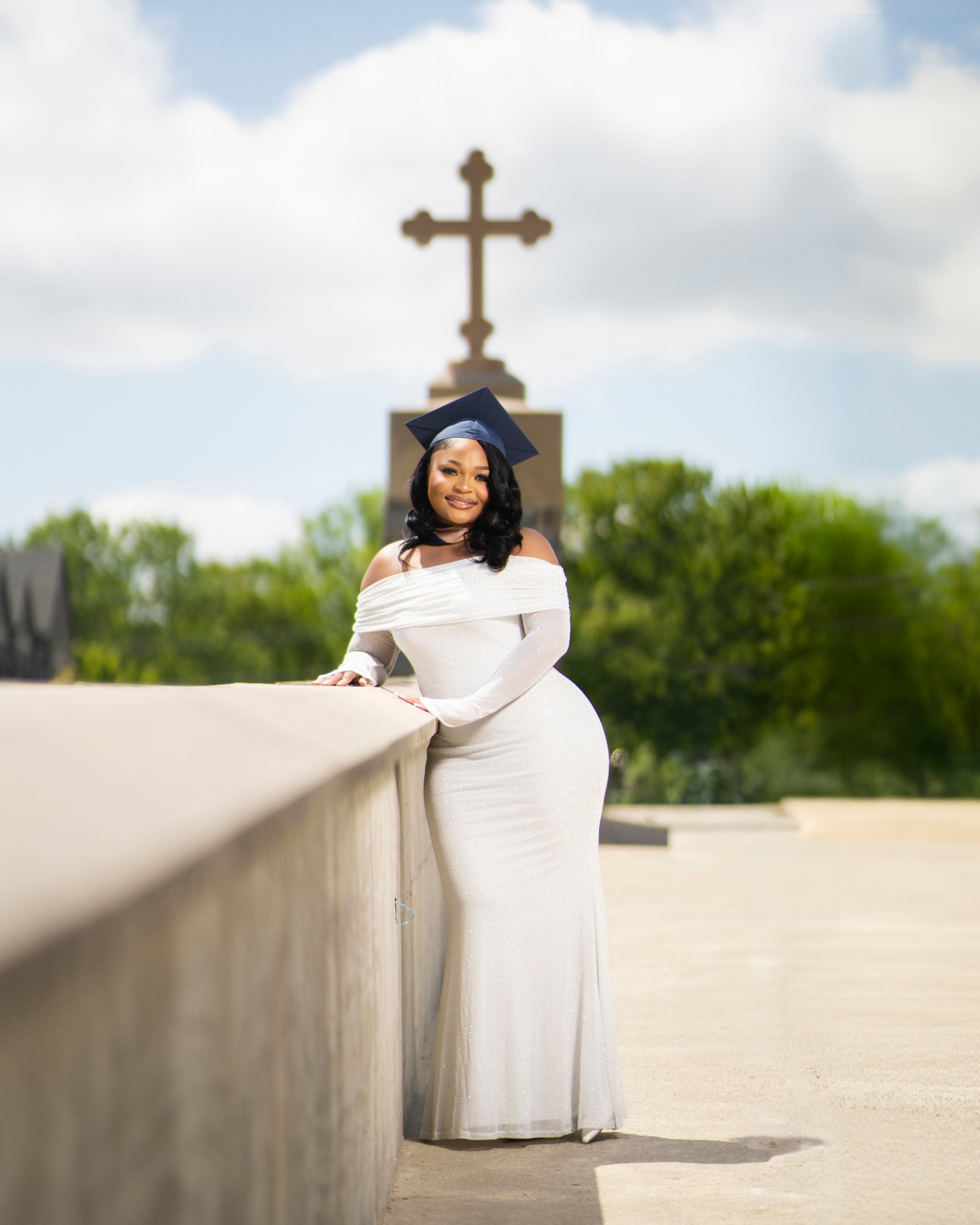 Grad in white gown by cross with blue sky backdrop
